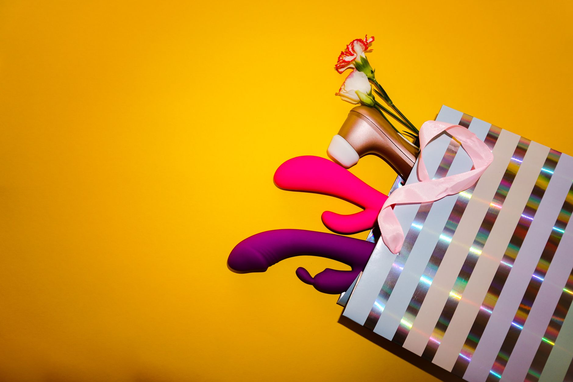 Colorful vibrators displayed in a holographic gift bag against a bold yellow background.
